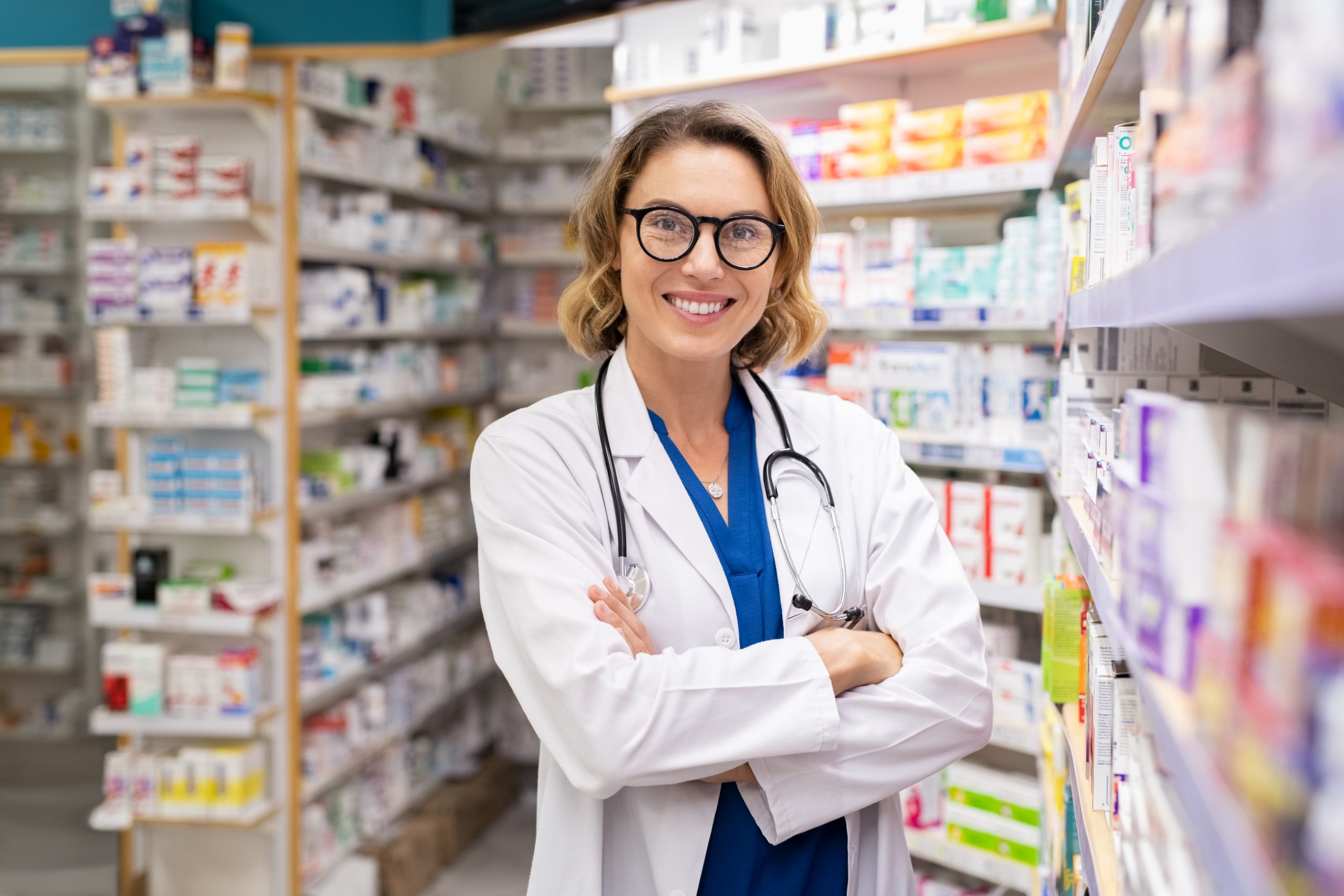 Portrait of mature woman pharmacist at pharmacy wearing labcoat with stethoscope. Happy smiling doctor standing in modern pharmacy drugstore. Friendly young pharmacist owner standing in shop and looking at camera.